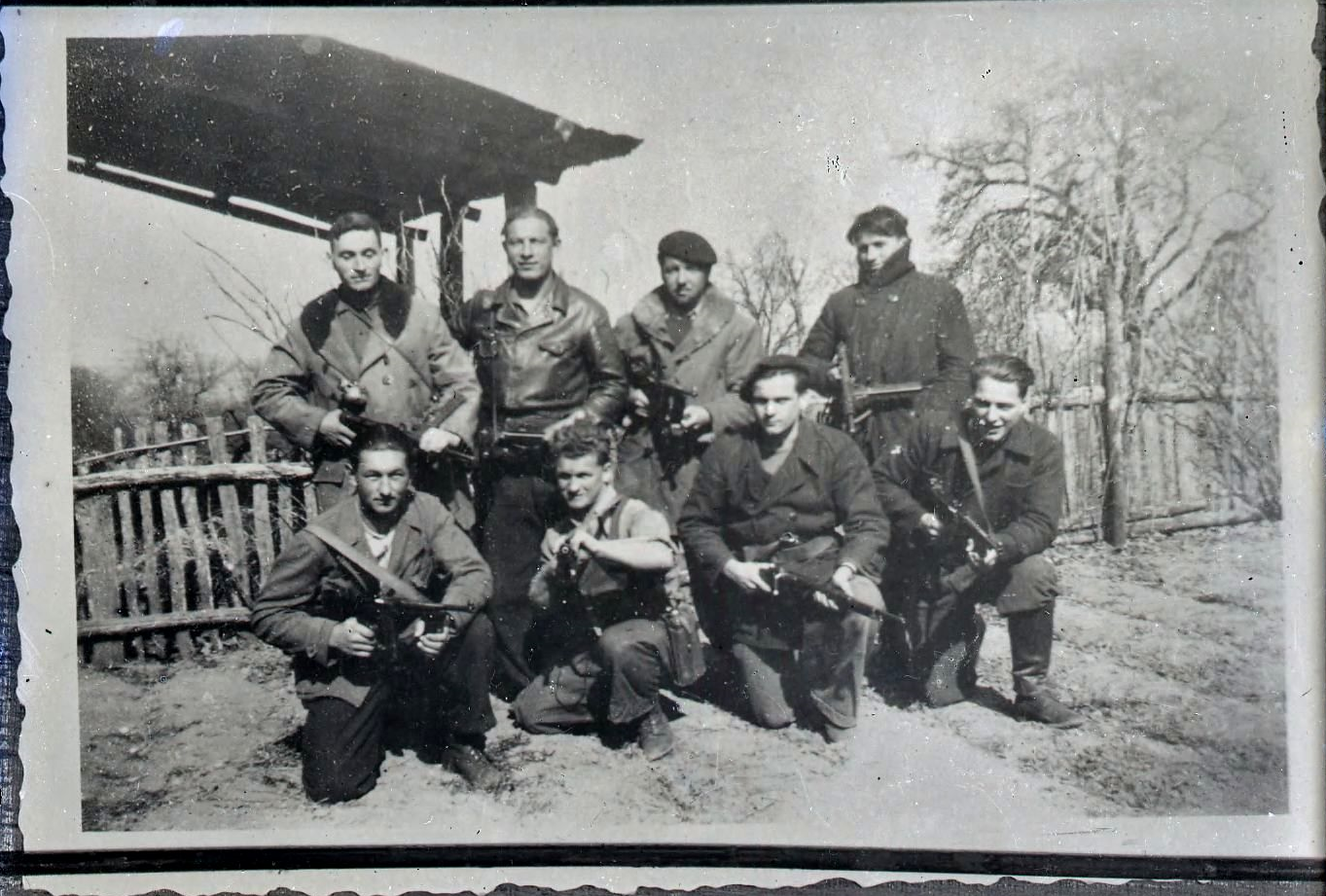 Photographie noir et blnac : un groupe de 8 jeunes hommes armés pose devant l'appareil ; derrière eux une haie et un arbre